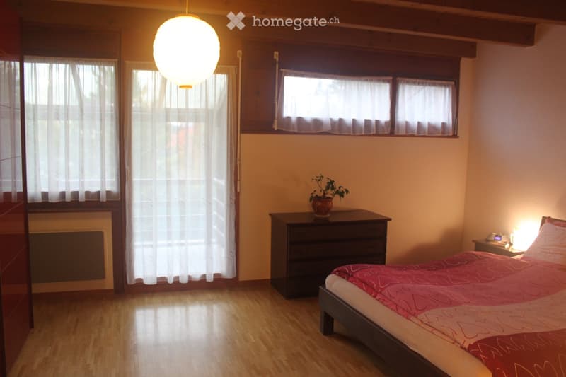 Cozy bedroom with wooden flooring, large windows with white curtains, a bed with red and white bedding, a nightstand with a lamp, a drawer unit, and a small potted plant on top.