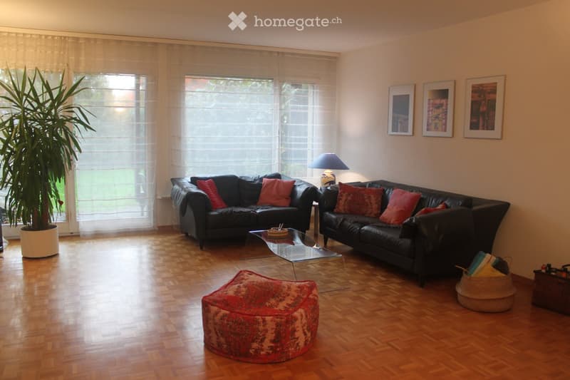 Living room with two black leather sofas, a glass coffee table, a red pouf, a floor lamp, and a large potted plant near the window.