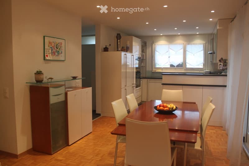 Modern kitchen and dining area with wooden flooring, white cabinets, a dining table with chairs, and a bowl of fruit on the table.