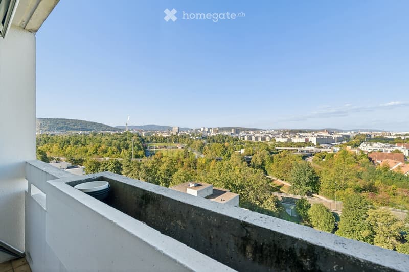 Balcony with a concrete railing, overlooking a cityscape with trees, buildings, and distant hills under a clear blue sky.