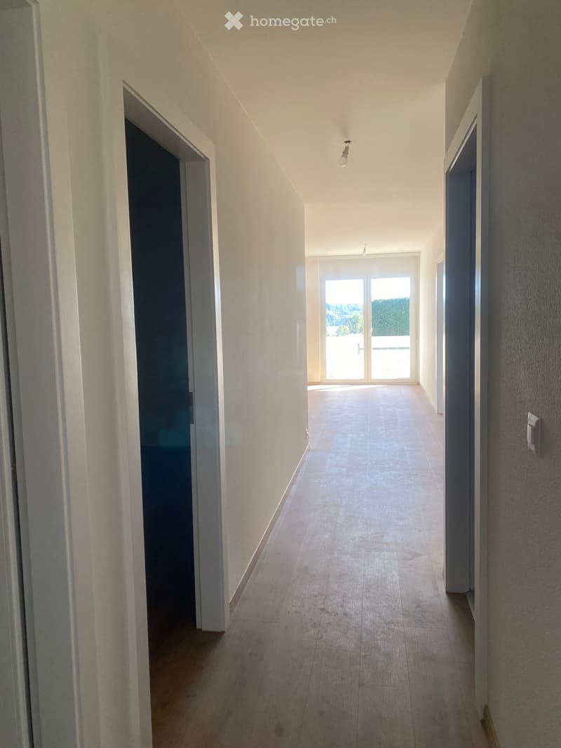 Empty hallway with light-colored walls, tiled floor, and recessed lighting. Features an open doorway leading to a balcony area with glass doors.