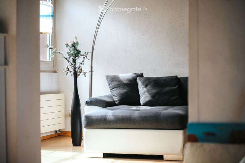 A modern living room with a grey sofa, a tall black vase with dried plants, and a sleek floor lamp near a window with blinds.