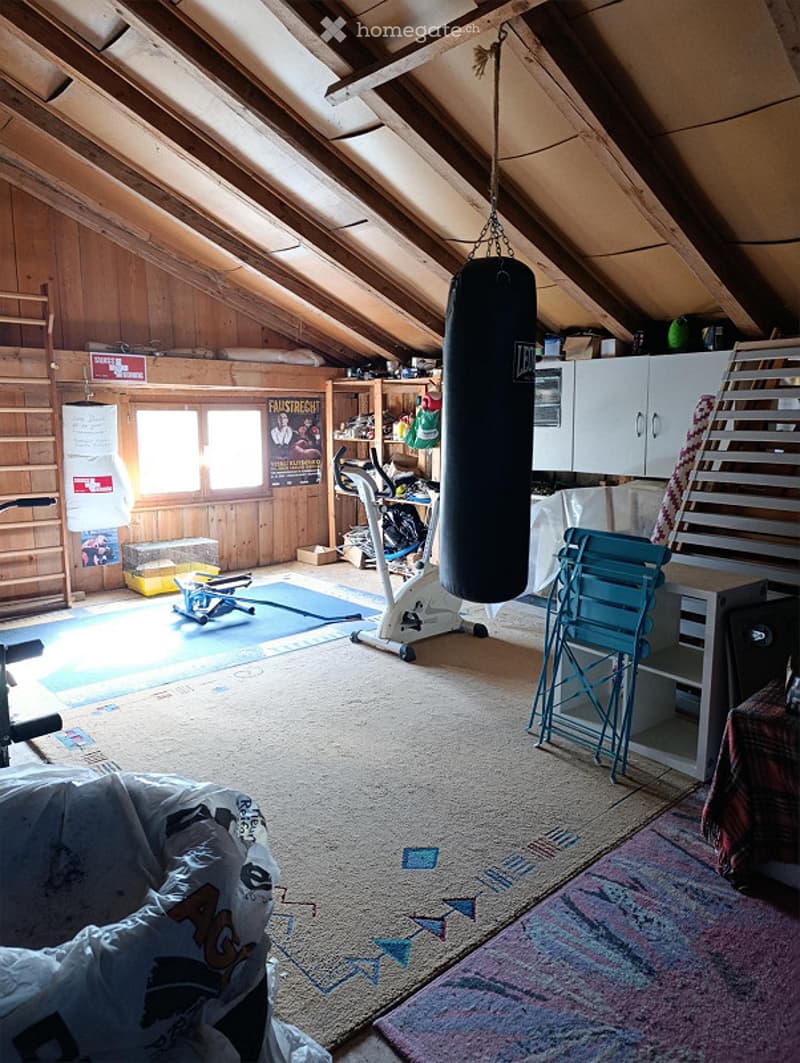 The room is an attic space with wooden beams and walls, featuring a punching bag hanging from the ceiling, an exercise bike, a chair, a small table with a laptop, a cabinet, shelves with various items, a poster on the wall, a stool, a bench, and a mirror.