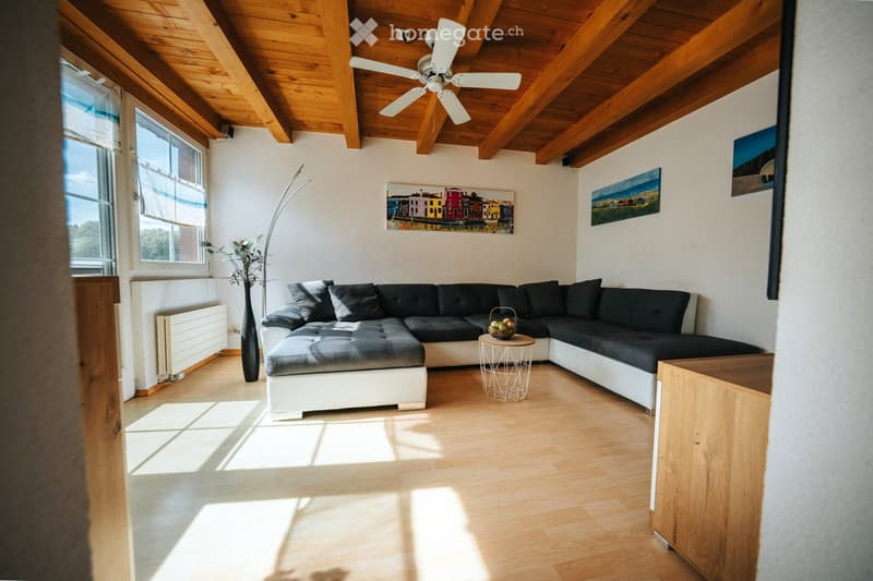 Modern living room with wooden ceiling beams, exposed wooden floor, large windows, grey sectional sofa, small round table, floor lamp, and artwork on walls.