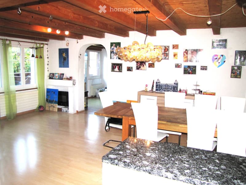 A well-lit living and dining area with wooden floors, a large wooden dining table surrounded by white chairs, a fireplace with a TV, and a balloon light fixture hanging from the ceiling. The walls are decorated with framed photos and artwork.