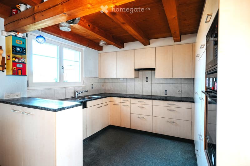 Bright kitchen with light-colored cabinets, a black countertop, and a sink. The room features a wooden ceiling with exposed beams and large windows allowing natural light to fill the space.