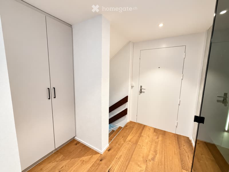 A clean, modern hallway with light wooden flooring, white walls, built-in white closet with black handles, and a closed white door leading to another room.