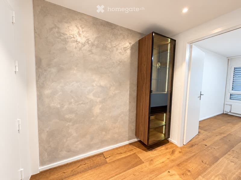 A modern, minimalist room with light wooden flooring, a textured beige wall, and a sleek wooden cabinet with glass doors.