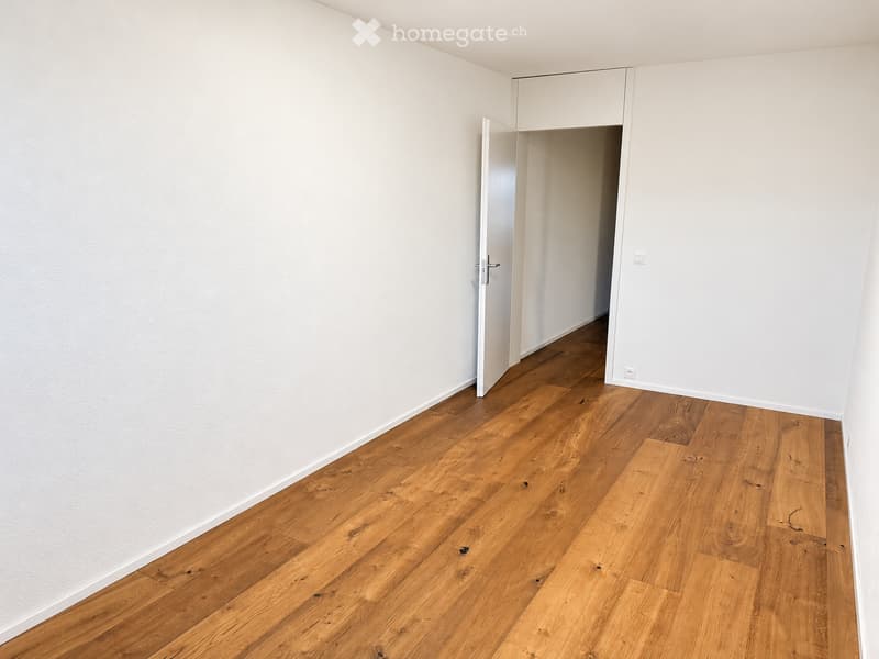 Empty room with light wooden flooring and white walls, featuring an open doorway leading to another room.