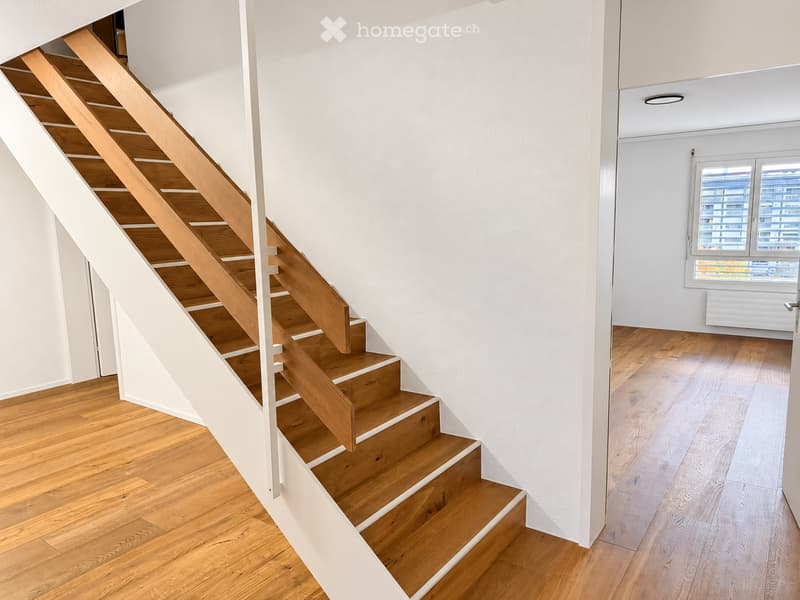 Modern hallway with wooden staircase, light-colored wooden flooring, white walls, and a window with blinds.