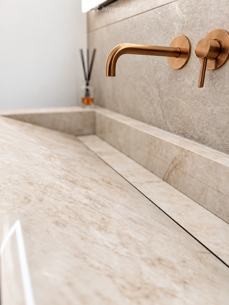 Modern bathroom sink with a sleek, gold-colored faucet mounted on a light beige marble countertop.
