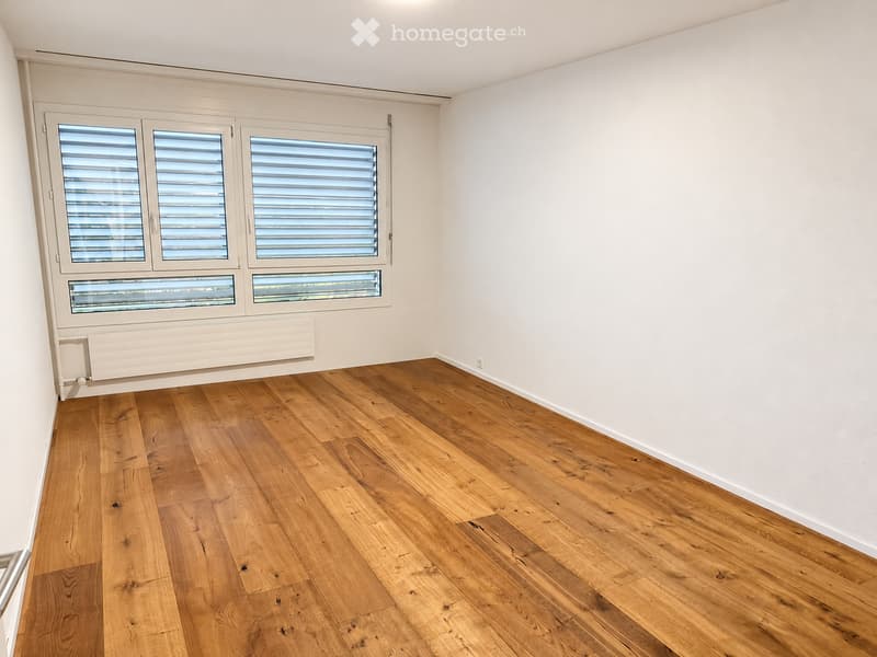 Empty room with light wooden flooring, white walls, and large windows with blinds. No furniture or equipment present.