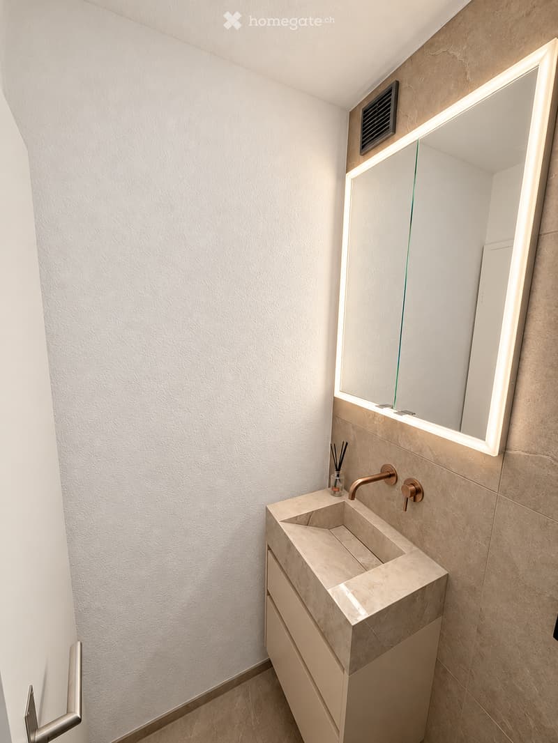 Modern bathroom with a sleek, minimalist design. Features a large, illuminated mirror, a stone sink with a modern faucet, and a built-in cabinet below.