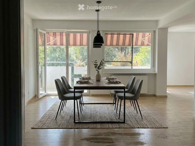 Modern dining room with a wooden dining table, six chairs, a vase with greenery, and tableware. Large windows with blinds and a view of greenery outside.