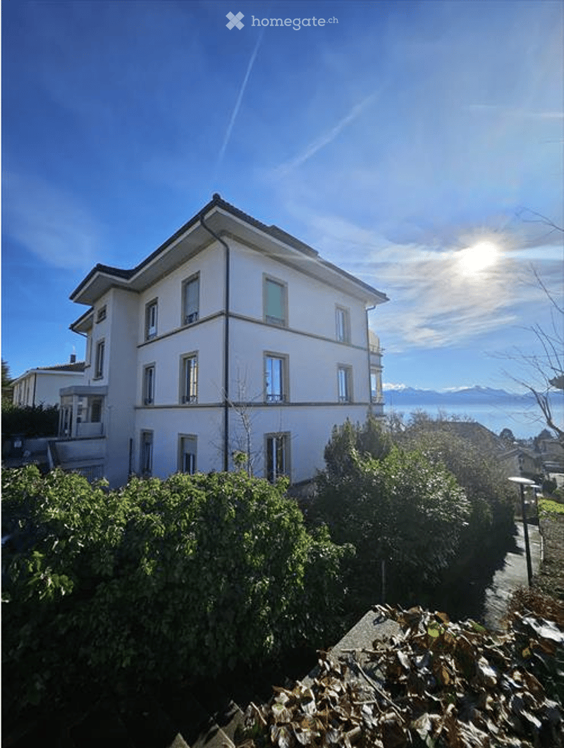 A multi-story residential building with white walls, multiple windows, and a view of mountains and a body of water in the background.