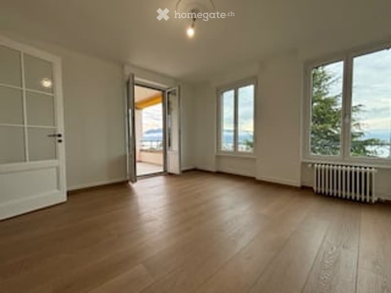 Bright, unfurnished room with wooden flooring, large windows offering a scenic view, and an open door leading to a balcony.
