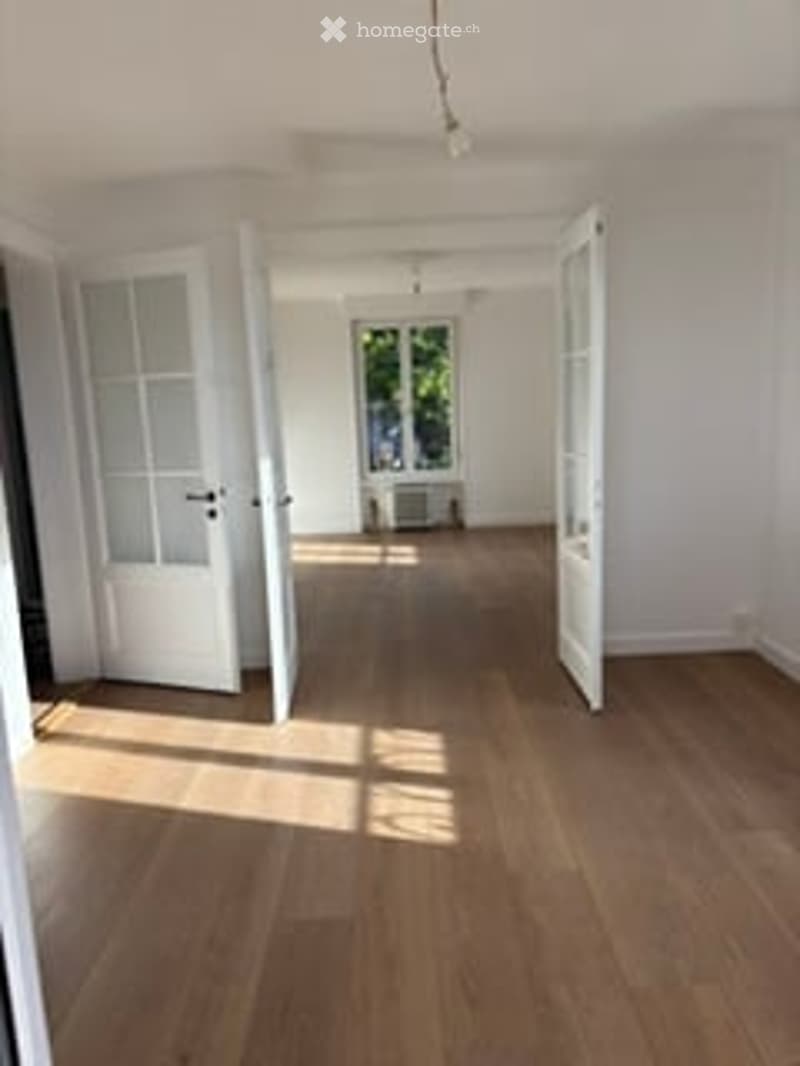 Empty room with wooden flooring, white walls, and large open doors leading to another space.