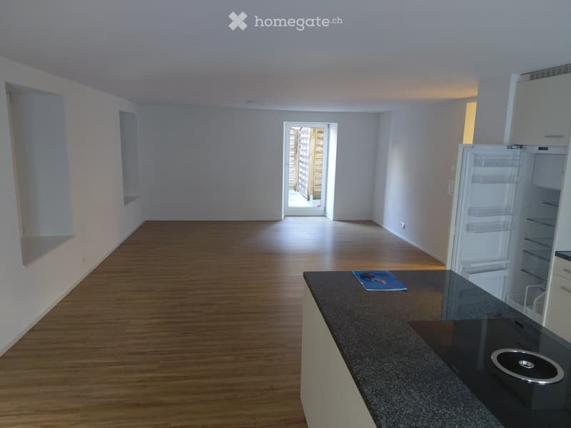 Empty room with wooden flooring, white walls, and a partially open door leading outside. A countertop with a small appliance and a few items is visible on the right side.