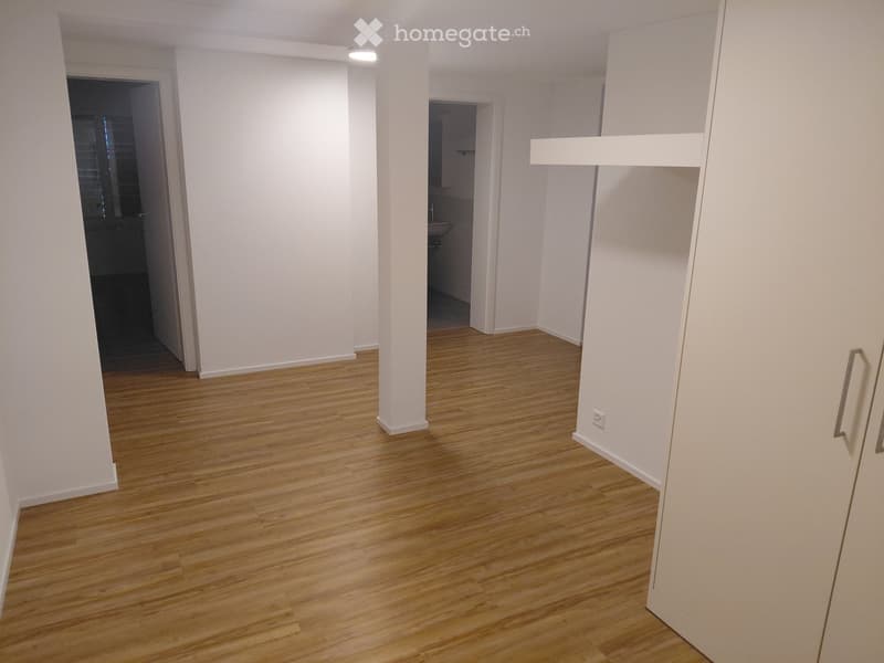 Empty room with wooden flooring and white walls, featuring a central column and doorways leading to other rooms.