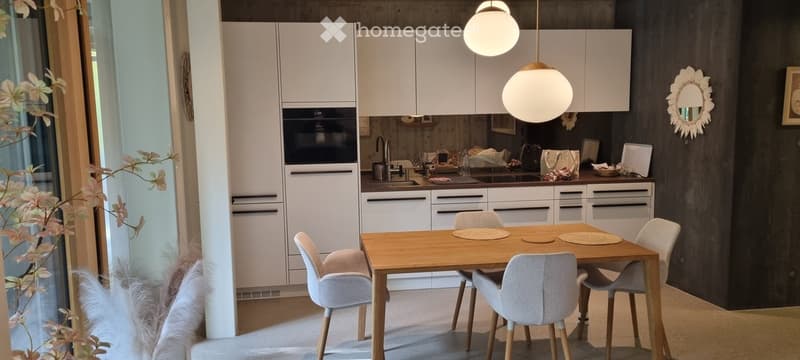 Modern kitchen and dining area with white cabinets, a wooden dining table with six chairs, pendant lights, and a sink area with an oven. Decorative items and a mirror are visible on the walls.