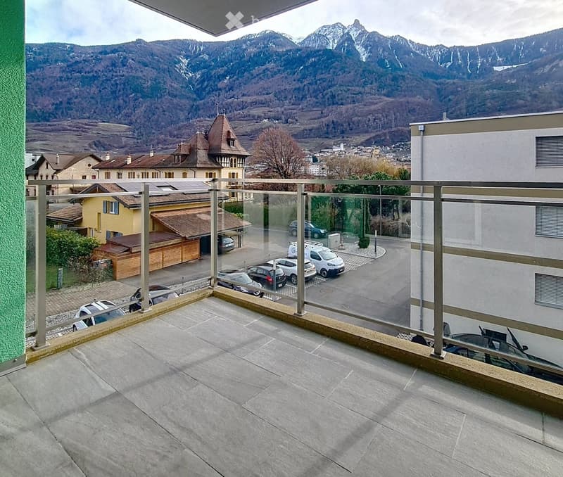 Balcony with a modern glass railing, overlooking a town and mountain range, with several parked cars visible below.