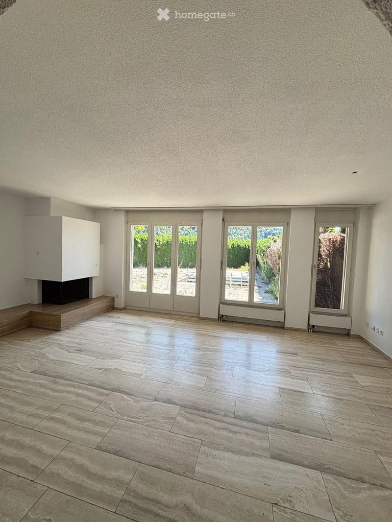 Empty room with light-colored tiled flooring, large glass doors leading to an outdoor area, and a built-in fireplace on the left side.