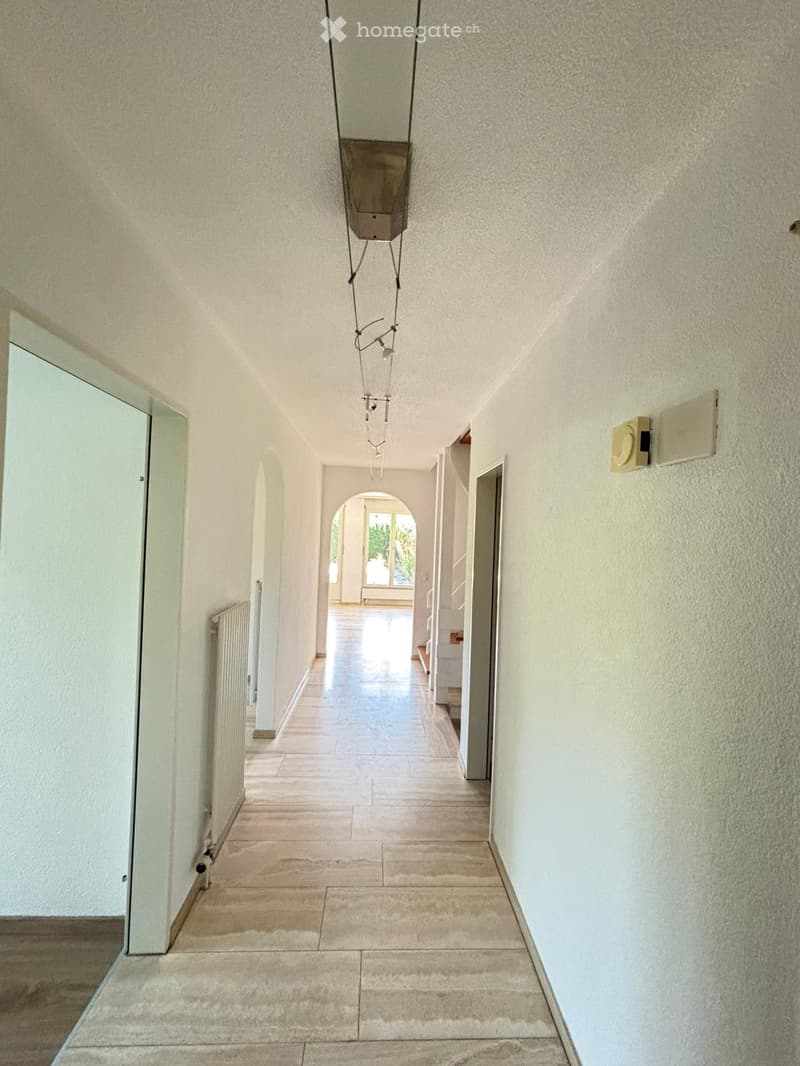A long, narrow hallway with beige tiled flooring and light-colored walls. Features include a hanging light fixture, a radiator on the left wall, and several doorways leading to other rooms.
