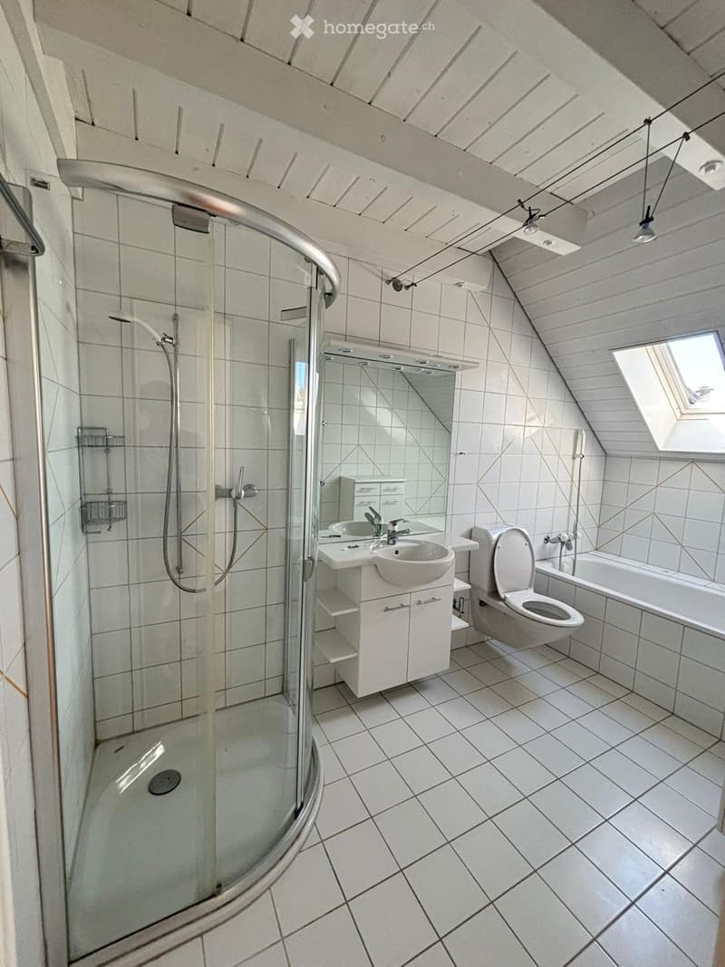 Modern bathroom with white tiled walls and floor, featuring a glass-enclosed shower, a sink with a mirror, a toilet, and a bathtub. The ceiling has exposed wooden beams and a skylight window.