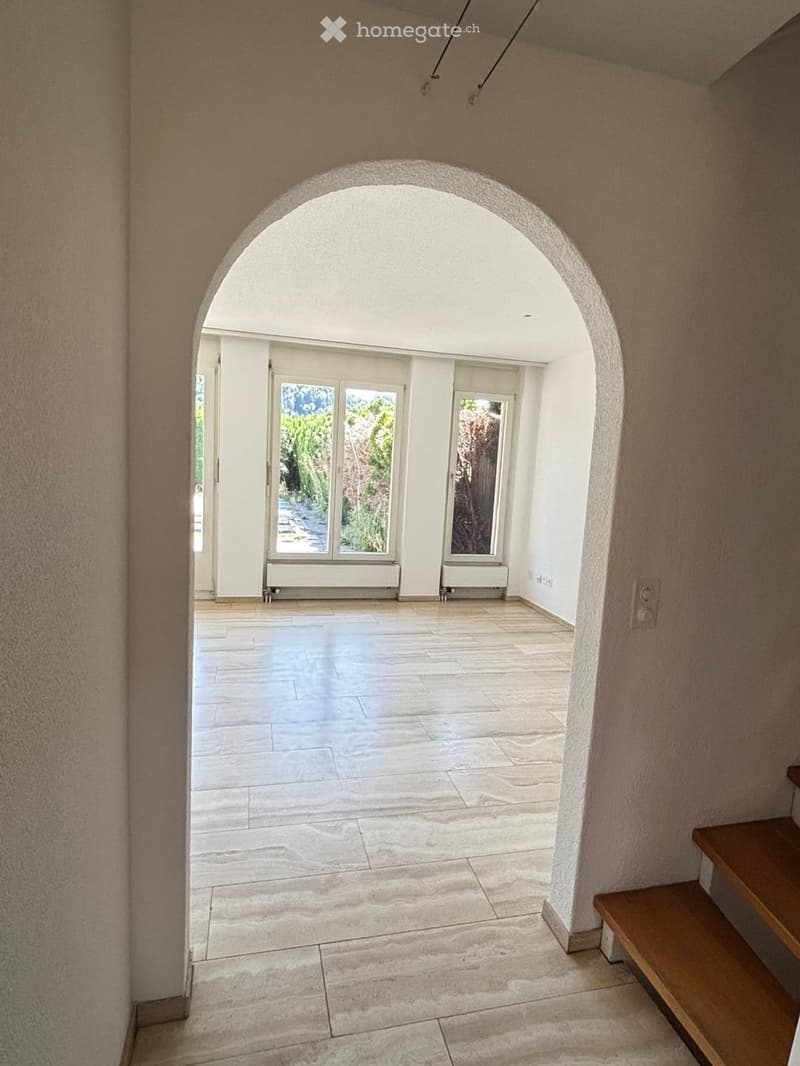 Empty hallway with beige walls, marble tiled floor, and an arched opening leading to a bright room with large windows and radiators.
