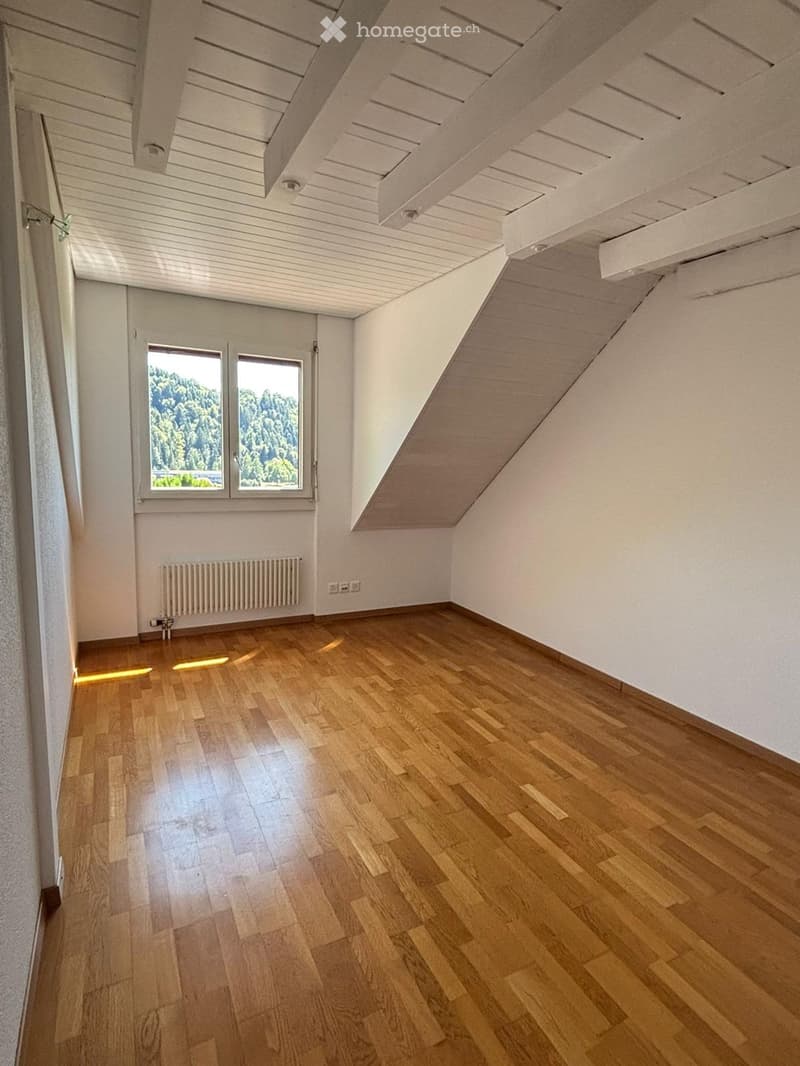 Empty room with wooden flooring, white walls, and a slanted ceiling. Features include a window with a view of greenery, a radiator, and a door.