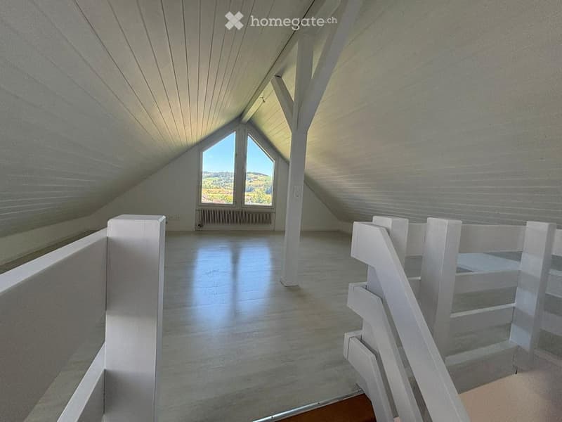 Bright, empty attic space with wooden floors, white walls, and large windows offering a scenic view. The room features a staircase leading upwards and built-in storage compartments along the walls.