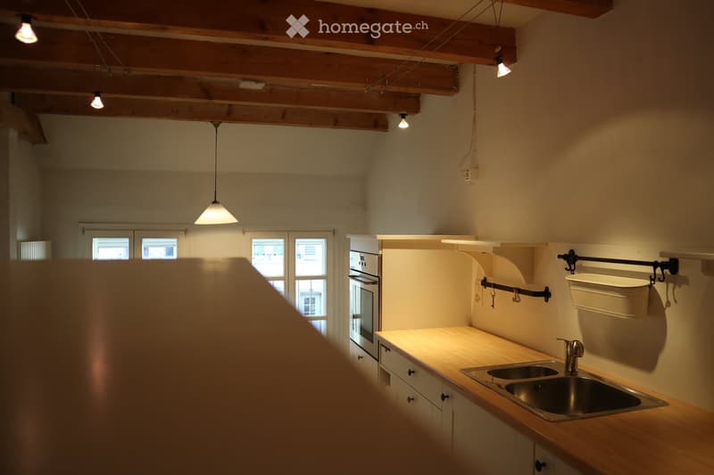Bright kitchen with wooden beams on the ceiling, pendant lights, a stainless steel sink, and white cabinets.