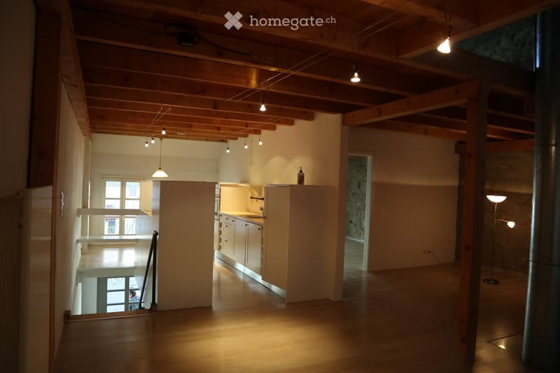 Open-plan living space with exposed wooden beams, a small kitchen area with cabinets and a sink, and a tall floor lamp in the corner.