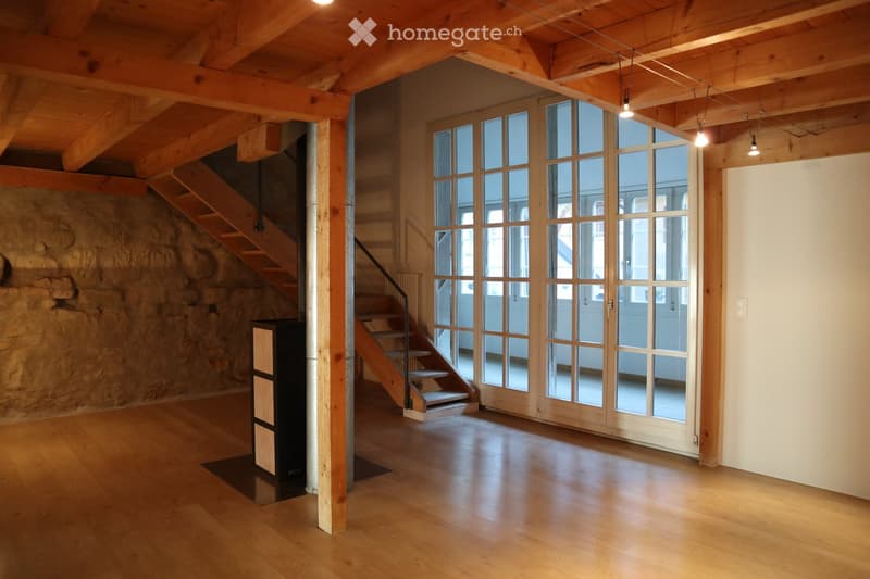 Empty room with wooden flooring, exposed wooden beams on the ceiling, large glass doors, and a wooden staircase leading upwards.
