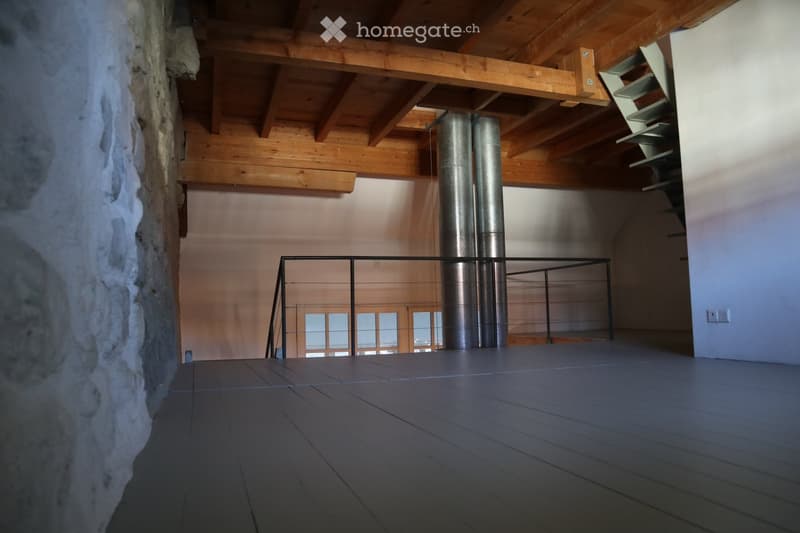 An empty room with wooden beams on the ceiling, a metal railing, and a stone wall. The floor is made of dark wood planks, and there is a large window in the background.