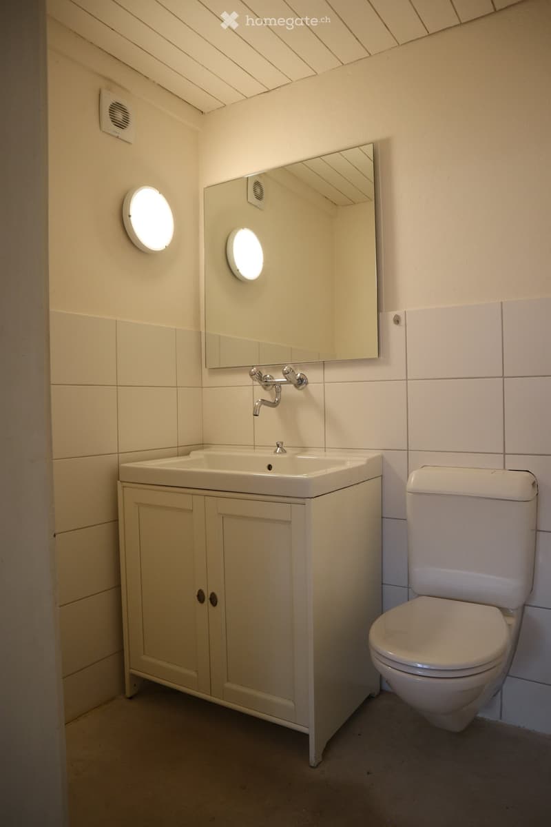 Modern bathroom with a white sink, toilet, and mirrored cabinet. Features round wall lights and tiled walls.