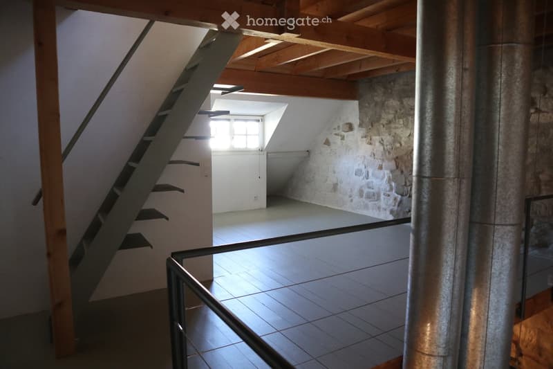 Empty room with exposed wooden beams, stone wall, tiled floor, and a staircase leading upwards.