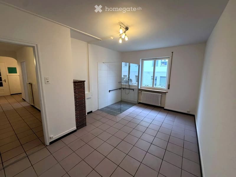Empty room with tiled floor, white walls, and a glass-enclosed shower area visible in the background. The room has a window with a radiator below it and a door leading to another area.