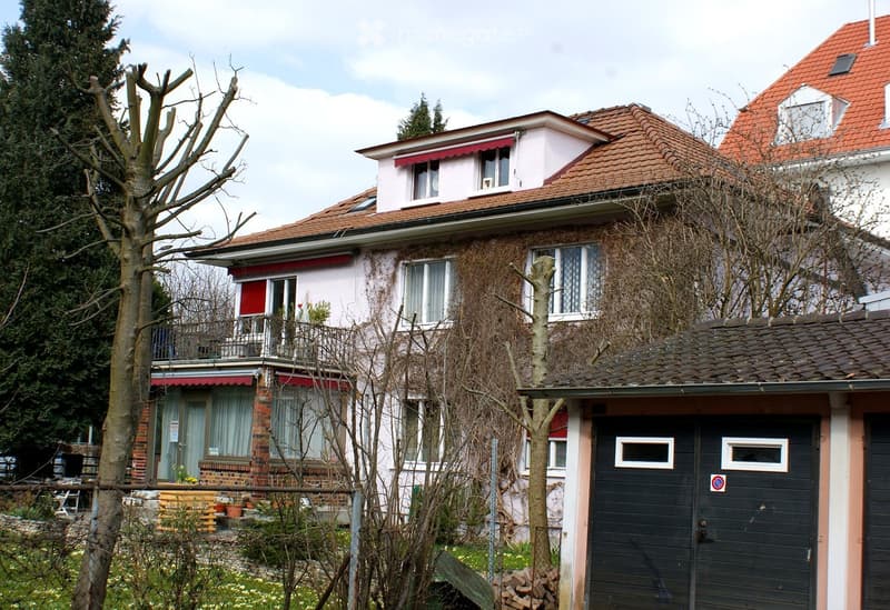 Two-story residential house with red-tiled roof, multiple windows, and a balcony on the upper floor. Features a garage with a black door and a small garden area with trees and potted plants.