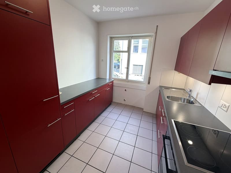 Modern kitchen with red cabinets, stainless steel sink, and electric stove. Features tiled flooring and large window with view of street.