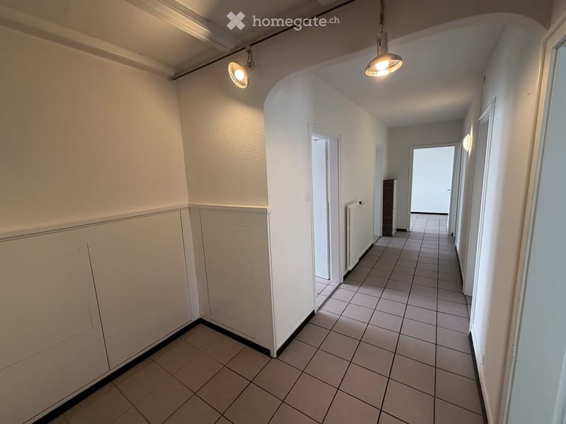 Empty hallway with tiled floor, white walls, and several doors leading to other rooms. The hallway is well-lit with ceiling lights and has a radiator on one side.