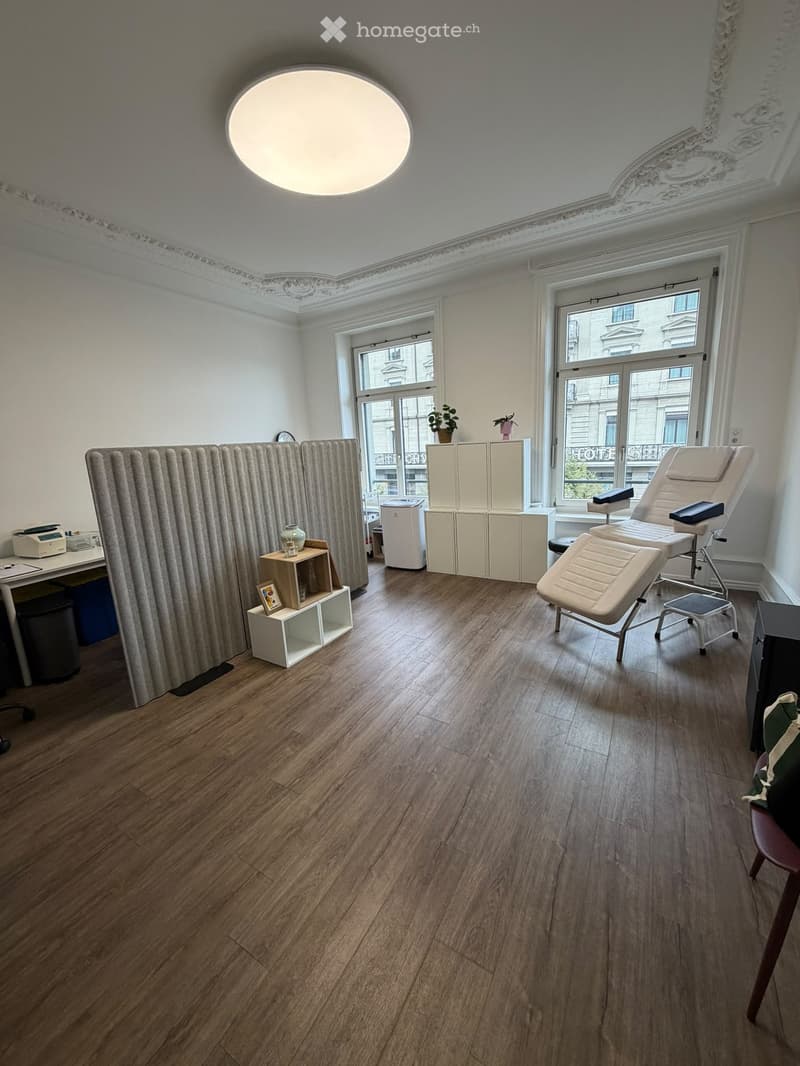 A well-lit room with wooden flooring, large windows, a massage chair, a small table with a vase, a cabinet, and a screen partition.