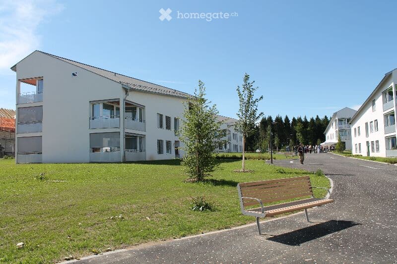 Modern white multi-story building with large windows and balconies, surrounded by green lawn and trees. A wooden bench is placed on the pathway in front of the building.