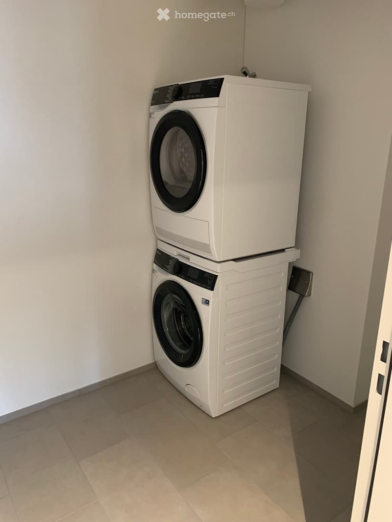 A small room with a stacked washing machine and dryer unit against a white wall, beige tiled floor.