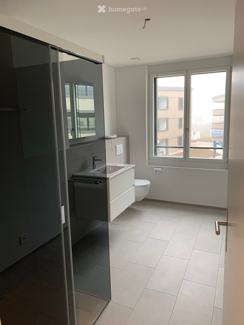 Modern bathroom with white tiled floor, dark grey cabinet, sink, toilet, and large window offering a view of the exterior.