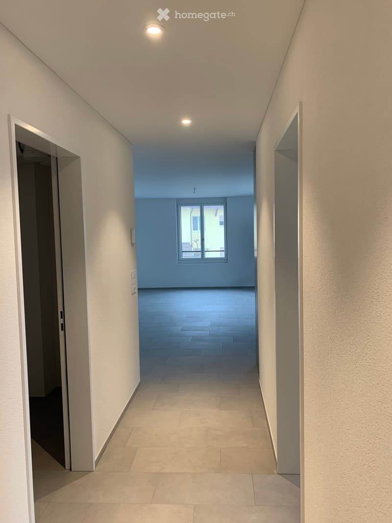 Clean, modern hallway with beige tiled flooring, white walls, and recessed lighting. Features two open doorways on either side and a window at the far end.