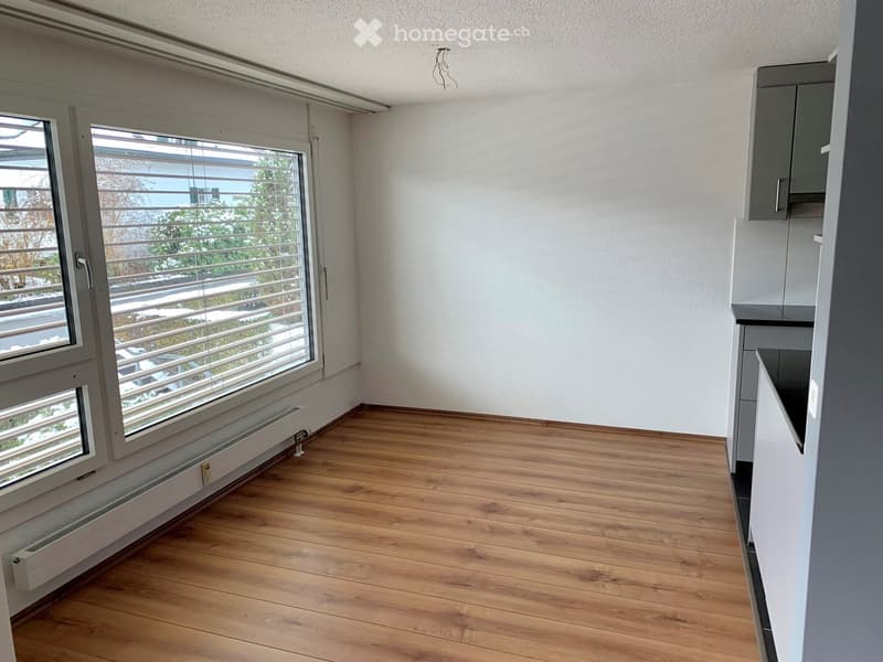 Empty room with large windows, wooden floor, white walls, radiator, and a modern kitchen design