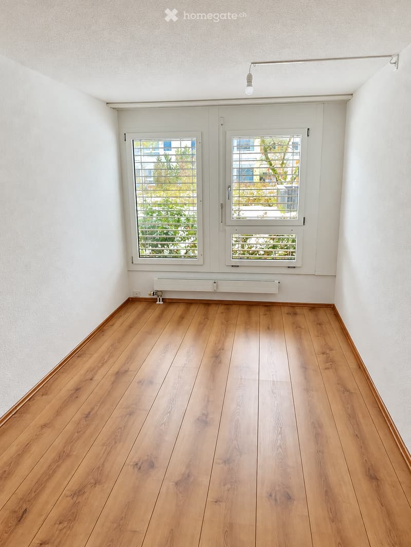 Empty room with wooden flooring, white walls, and two windows with blinds. A radiator is mounted on the wall below the windows.