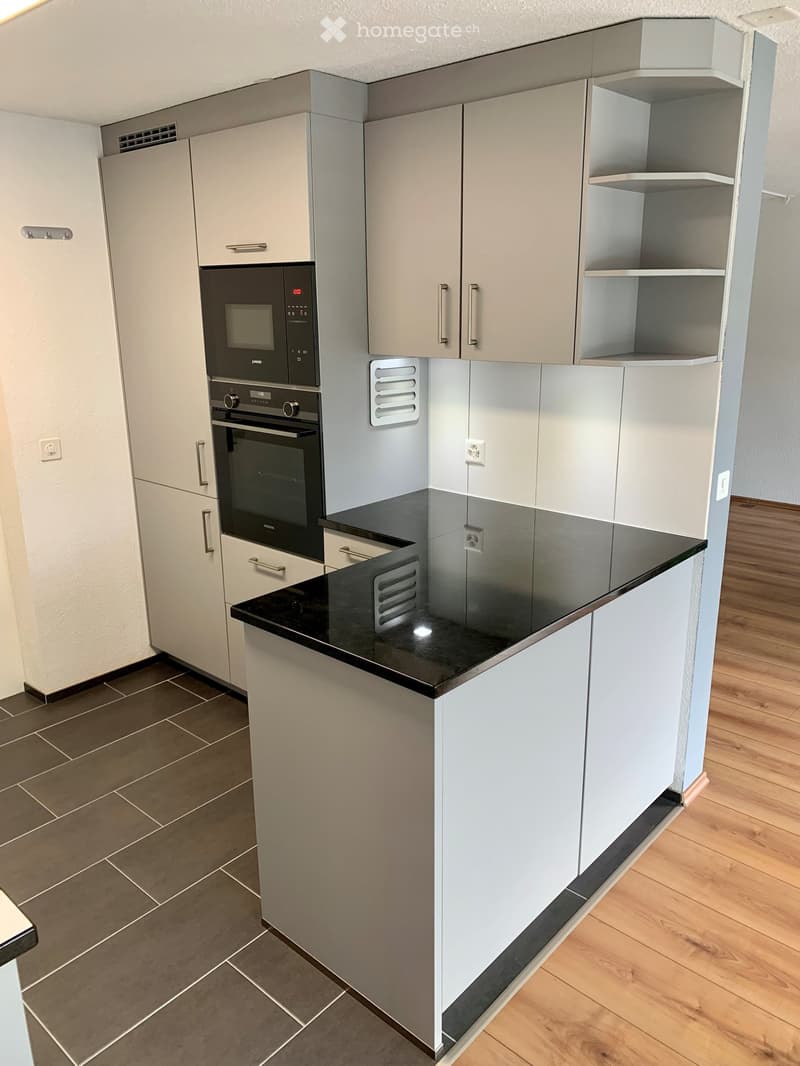 Modern kitchen with wooden floor, built-in oven and microwave, large central island, gray and white cabinetry, granite countertop, tiled backsplash.