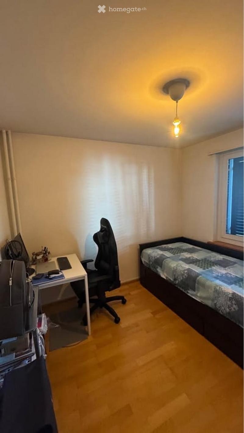 Cozy bedroom with a single bed, desk with a laptop, and a chair. The room features wooden flooring and a window with blinds.